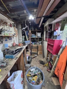 Cluttered wooden storage shed or workshop with tools, shelves, a workbench, and stored equipment.