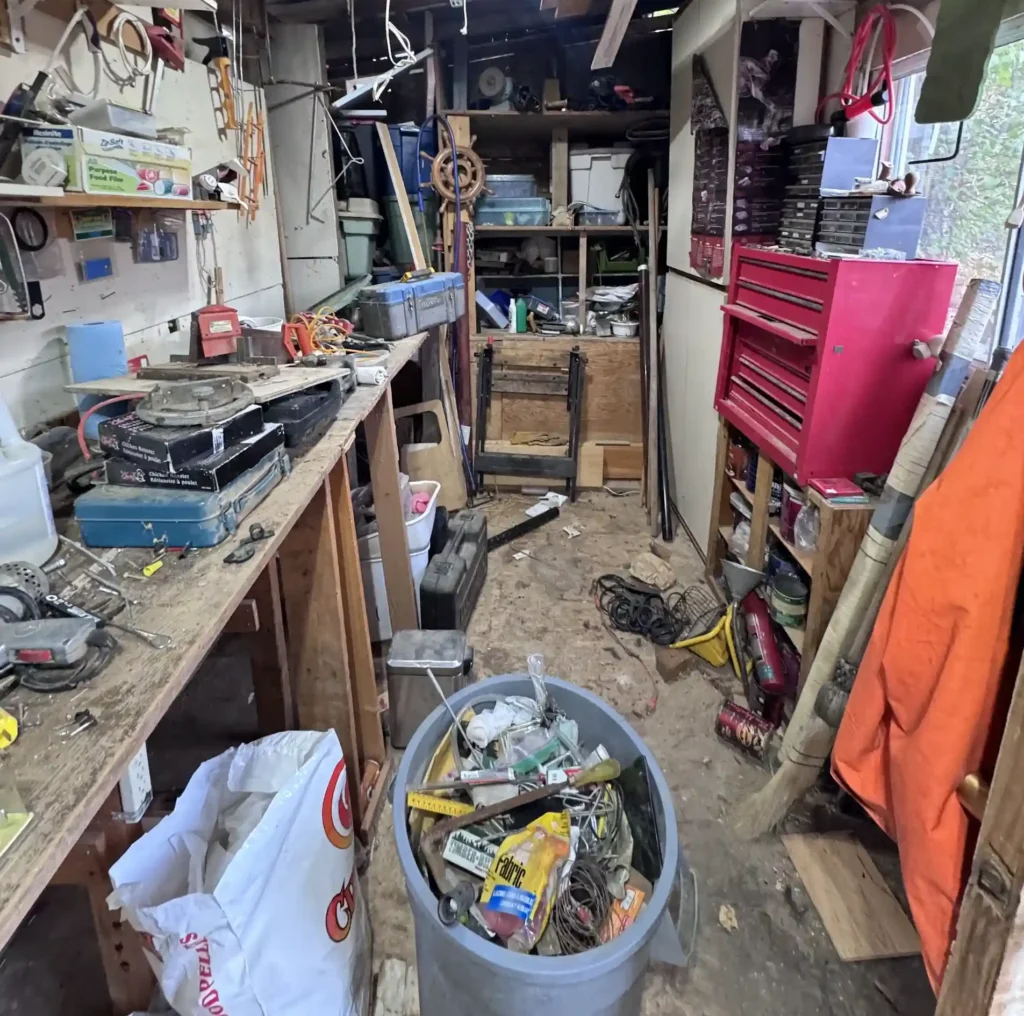 Cluttered wooden storage shed or workshop with tools, shelves, a workbench, and stored equipment.